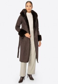 Brązowy płaszcz z sztucznego zamszu z puszystym brązowym futrzanym kołnierzem i mankietami, z zapięciem na guziki i prostym krojem. Połączony z jasnymi spodniami.