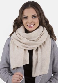 Beige huivi, joka on valmistettu pehmeästä villasta ja jossa on paksu, mukava rakenne. Se laskeutuu löyhänä kaulan ympäri, täydentäen harmaata takkia.