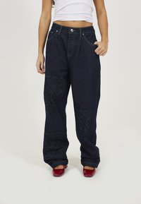 Dark blue denim wide-leg pants with green stitching and oversized logo patterns, featuring front pockets and a button closure.