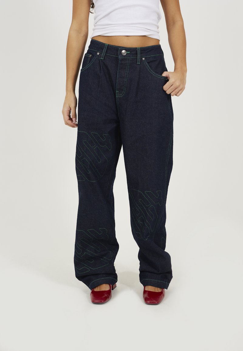 Dark blue denim wide-leg pants with green stitching and oversized logo patterns, featuring front pockets and a button closure.