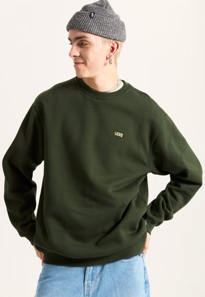 Olive green sweatshirt with a crew neck, ribbed cuffs, and a small gold Vans logo. Worn over a light blue shirt with denim shorts.