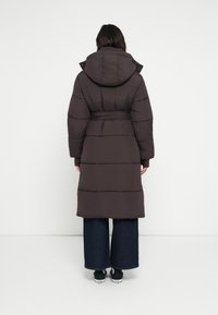 Καφέ επενδυμένο παλτό με κουκούλα, ζώνη στη μέση και οριζόντια ραφή. Φοριέται πάνω από φαρδιά σκουρόχρωμα τζιν παντελόνια και μαύρα παπούτσια.