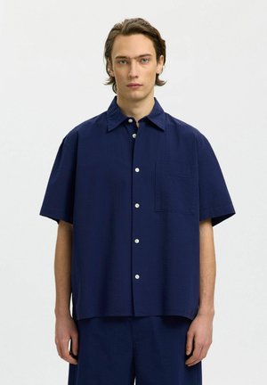 Młody mężczyzna o średniej długości włosach, ubrany w ciemnogranatową koszulę na krótkie rękawy zapinaną na guziki oraz pasujące spodnie na jednolitym białym tle.