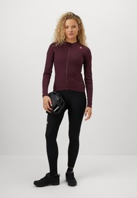 Bordeauxfarbendes langärmeliges Radtrikot mit einem Reißverschluss vorne, kombiniert mit schwarzen, figurbetonten Leggings. Das Model hält einen schwarzen Helm in der Hand.