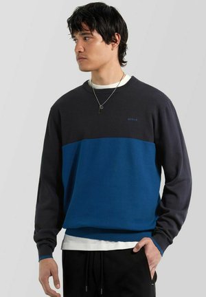 Joven con jersey a bloques de color negro y azul, camisa blanca debajo, pantalones negros y un collar plateado, mirando hacia la izquierda.