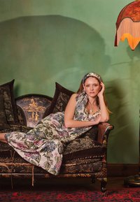 Abito maxi con motivo floreale in tessuto leggero, con tonalità di rosa e verde. Divano ornato scuro con cuscini decorativi e lampada vintage.