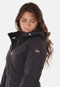 Giacca nera impermeabile con cappuccio, chiusura a zip sul davanti, tasche laterali e patch con logo dorato. Realizzata in tessuto morbido con un design aderente.