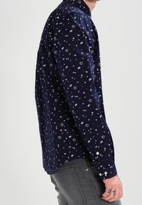 Tmavě modrá košile s dlouhým rukávem s rozptýleným bílým vzorem malých tvarů. Manžety na knoflíky a klasický límec. Hladká textura.