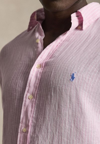 Mees kannab heleroosat, vertikaalselt triibulist nööbitavat särki sinise tikitud polo mängija logoga rinnal.