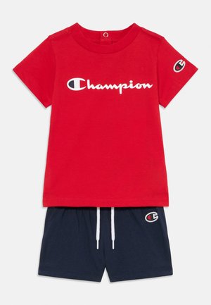 T-shirt rouge à manches courtes avec logo Champion blanc et short bleu marine avec cordons blancs et logo Champion sur la jambe gauche.