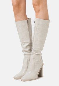 Bottes hauteur genou en faux cuir gris clair avec une texture crocodile, présentant une fermeture éclair latérale et un talon bloc épais.