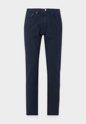 Jeune homme portant une chemise blanche à boutons, un pantalon bleu foncé et des mocassins marron foncé, debout avec une main dans la poche sur fond blanc.