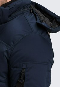 Navyblauwe pufferjack met een hoge kraag, ritsen op de borst en schouder, en een textuurstof. Bevat een zijzak met hardwareaccenten.