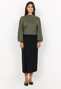 Olivgrünes Langarmoberteil mit Ballonärmeln, kombiniert mit einem taillierten schwarzen Midirock mit seitlichem Schlitz. Schwarze Ballerinas runden das Outfit ab.