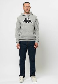 Grijze pullover hoodie met zwart logo op de borst, voorzak, ribgebreide manchetten, gecombineerd met donkerblauwe denim jeans en witte sneakers.