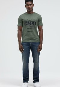 Man wearing an olive green graphic t-shirt, blue jeans, and white sneakers, standing against a plain light gray background.