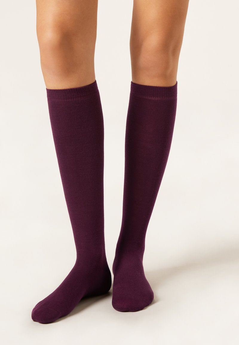 Jambes portant des chaussettes hautes violet foncé, debout sur une surface claire avec un pied à plat et l'autre légèrement relevé.