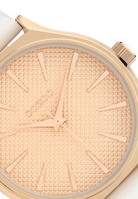 Gold-tone watch with a textured beige dial, elongated hour markers, and slender hands. Features a white strap and a crown on the side.