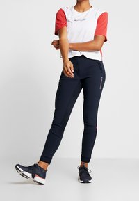 Osoba oblečená ve světlém a červeném tričku s krátkým rukávem, v námořnických sportovních kalhotách a námořnických sportovních botách, stojící s jednou nohou mírně zvednutou.