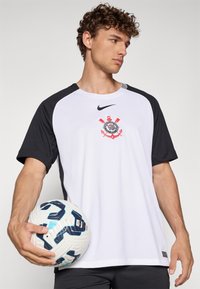 Nike t-skjorte med hvitt kropp og svarte ermer, brodert logo og rund hals. Modellen holder en mønstret fotball.