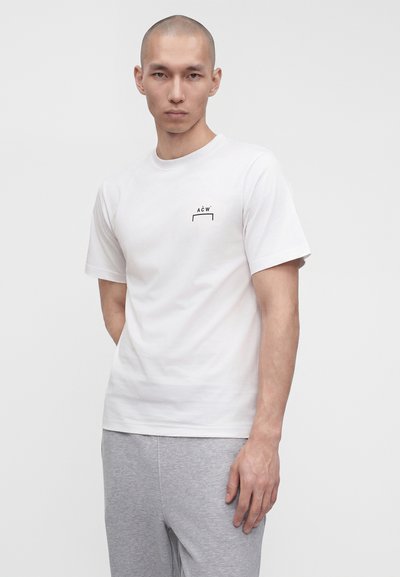 Άνδρας με κοντά μαλλιά που φοράει λευκό T-shirt με μικρό μαύρο λογότυπο "A C W" στο στήθος και ανοιχτό γκρι παντελόνι, στέκεται μπροστά σε απλό φόντο.