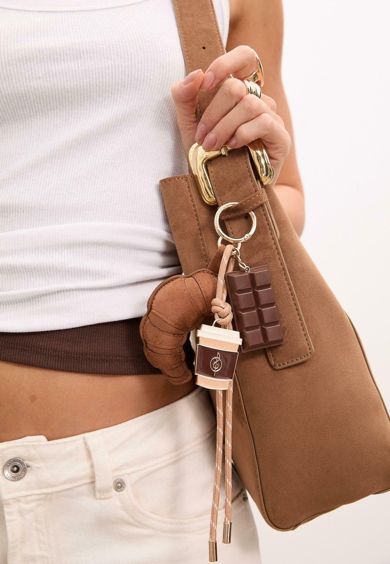 Une personne tient un sac brun avec un fermoir doré, comportant des porte-clés en forme de croissant, de barre de chocolat et de tasse de café sur un fond clair.