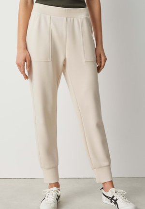 Femme portant un pantalon de jogging couleur crème avec de grandes poches avant et des baskets blanches à rayures noires, se tenant devant un fond uni.