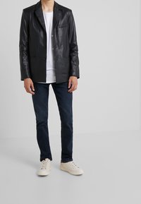 Blazer en cuir noir avec une poche unique, associé à un t-shirt blanc, un jean bleu et des baskets blanches. Texture lisse et design ajusté.