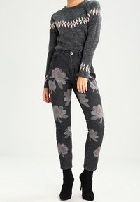 Suéter gris con estampado, de manga larga, combinado con jeans negros que presentan grandes estampados florales en rosa apagado y gris; acompañado de botas negras hasta el tobillo.