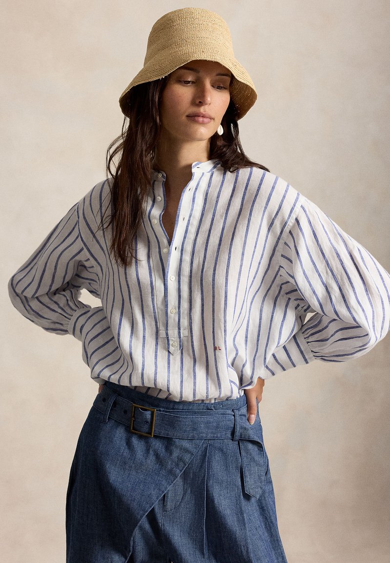 Kvinne med langt, mørkt hår iført beige bøttehatt, hvit skjorte med blå vertikale striper, og høytlivs blå skjørt med belte, hendene på hoftene.