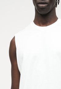 Κοντινό πλάνο ενός ατόμου που φοράει λευκό αμάνικο πουκάμισο, εμφανίζοντας τον λαιμό, τα χείλη και το πάνω μέρος του κορμού μπροστά σε ένα απλό φωτεινό φόντο.