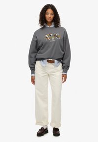 Sudadera gris de gran tamaño con bordado floral, superpuesta sobre una camisa a rayas azul, combinada con pantalones de pierna ancha color crema y mocasines oscuros.
