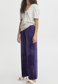 Migotający srebrny top z cekinami w kształcie litery V, zestawiony z fioletowymi welurowymi spodniami o szerokich nogawkach oraz beżowymi, odkrytymi szpilkami. Gładka tekstura, luźny krój.