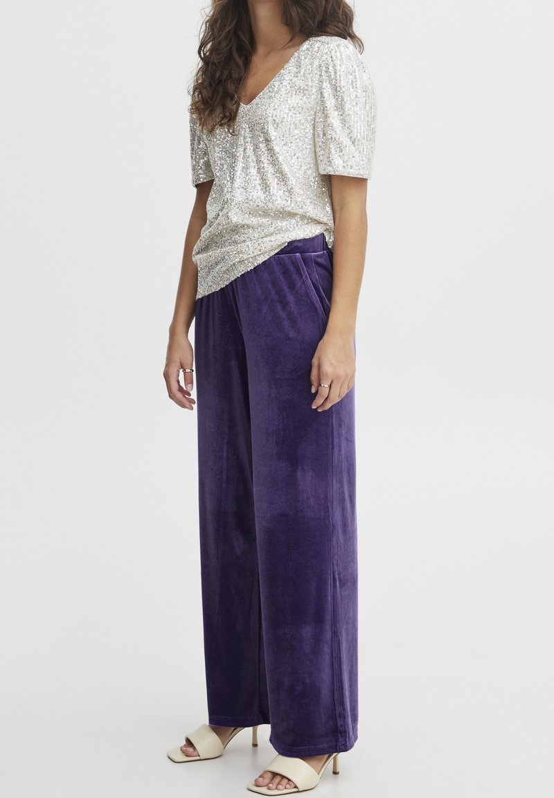 Migotający srebrny top z cekinami w kształcie litery V, zestawiony z fioletowymi welurowymi spodniami o szerokich nogawkach oraz beżowymi, odkrytymi szpilkami. Gładka tekstura, luźny krój.