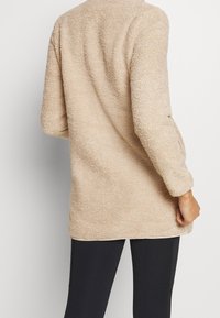 Beżowy polarowy sweter z długimi rękawami, o miękkiej strukturze i zaokrąglonym dolnym wykończeniem, noszony na czarnych legginsach. Brak widocznych wzorów lub elementów metalowych.