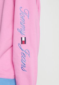 Felpa rosa con accenti blu, con la scritta "Tommy Jeans" ricamata in corsivo lungo la manica e una piccola toppa del logo.
