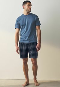 Světle modré bavlněné tričko v kombinaci s tmavými kostkovanými šortkami s zelenými a šedými akcenty. Model stojí bosý před neutrálním pozadím.