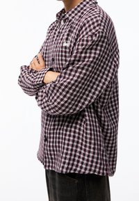 Persona che indossa una camicia a maniche lunghe a scacchi bordeaux e bianca con le braccia incrociate, abbinata a pantaloni scuri, in piedi di lato.