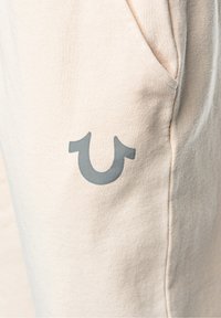 Beige cotton pants featuring a side pocket and a prominent gray logo resembling a stylized horseshoe on the left thigh.