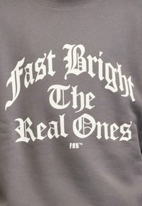Grå hettegenser med fet hvit tekst: "Fast Bright The Real Ones" i gotisk skrift. Mykt stoff med rund hals og ribbestrikkede mansjetter.