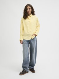 Lys gul skjorte med krage, kombinert med falmede blå vide bukser og mørkebrune snøringsko. Glatt stoffstruktur.