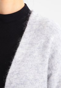 Ljust grått ullkardigan med öppen front, mjuk textur och en mysig känsla, tillsammans med en svart ribbad polotröja under.