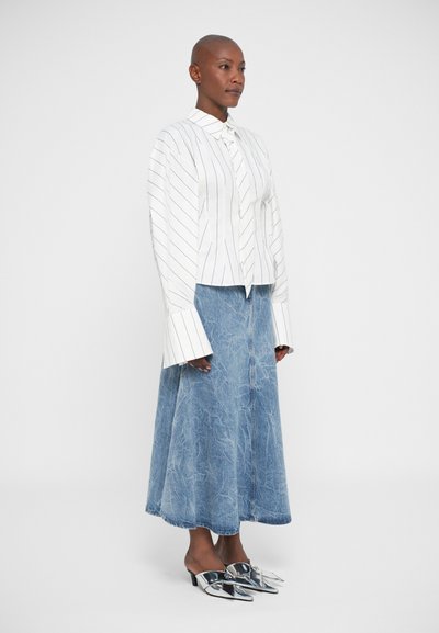 Woman standing wearing a white striped long-sleeve blouse with extended cuffs, a light blue denim midi skirt, and silver pointed heels.