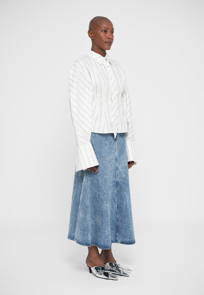 Eine Frau steht und trägt eine weiß gestreifte Bluse mit langen Ärmeln und verlängerten Manschetten, einen hellblauen Jeans-Midirock und silberne spitze High Heels.