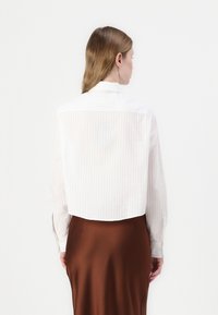 Camicia bianca tagliata con righe verticali, maniche lunghe e colletto classico, abbinata a una gonna di raso marrone.