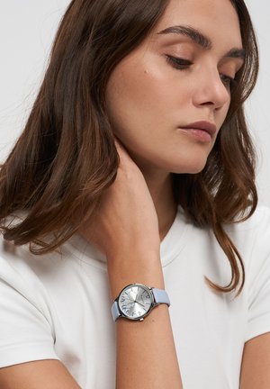 Frau mit weißem Hemd und mittelbraunen Haaren trägt eine silberne Uhr mit hellblauem Armband, legt die Hand sanft an ihren Hals.