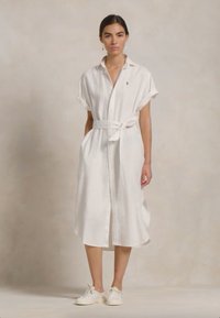 Robe chemise en lin blanc avec des manches courtes retroussées et une ceinture à la taille. Présente un devant à boutons et une coupe décontractée. Portée avec des baskets blanches.