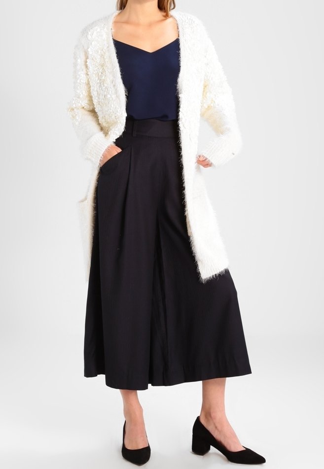 Een lange, fluffy witte cardigan over een marineblauw topje, in combinatie met hoge, zwarte culottes. Zwarte puntige hakken maken de outfit compleet.