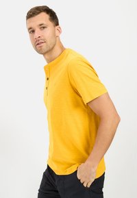 Kortærmet gul henley skjorte med tre knapper og rund udskæring, lavet af blødt materiale, parret med mørke shorts.