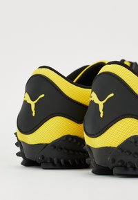 Paire de chaussures de sport Puma jaunes et noires vues de dos, montrant des semelles texturées et le logo Puma sur les contreforts des talons.